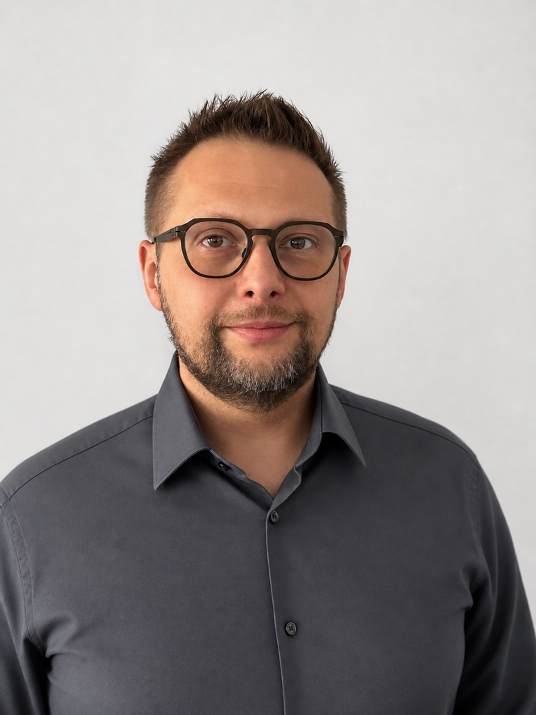 Na zdjęciu widzimy mężczyznę w średnim wieku, stojącego na tle jasnego, jednolitego tła. Ma krótkie, lekko przyciemniane włosy i niewielką brodę. Ubrany jest w elegancką, szarą koszulę z długimi rękawami. Spogląda na widza, jego twarz wyraża neutralny, lecz pewny siebie wyraz. Oczy ma koloru jasnobrązowego, a jego spojrzenie jest bezpośrednie. Koszula jest starannie wyprasowana, co dodaje elegancji jego wyglądowi.